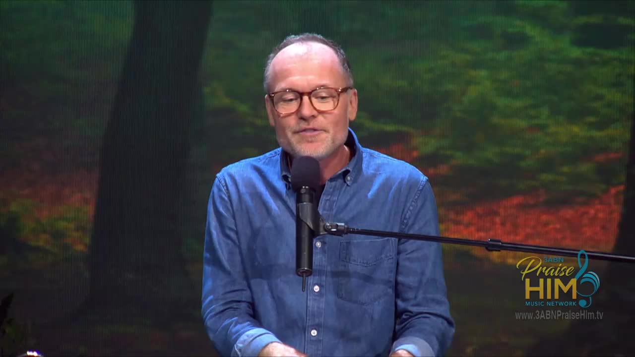 A man in a denim shirt speaks into a microphone, framed against a backdrop that suggests a forest. The 3ABN Praise Him Music Network logo is visible on the lower right.
