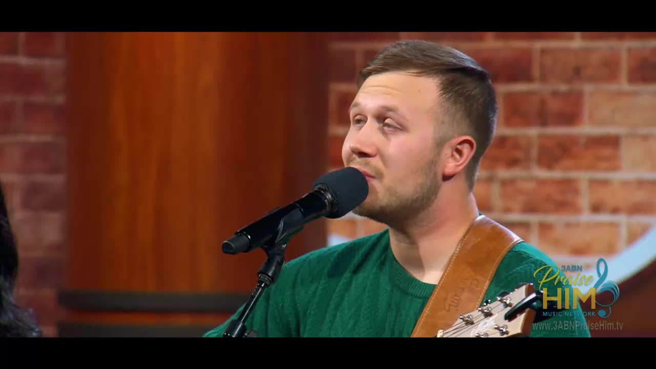 On a stage in the United States, a man in a green sweater sings into a microphone, his guitar strap visible over his shoulder. The 3ABN Praise Him Music Network logo is in the corner as he performs.
