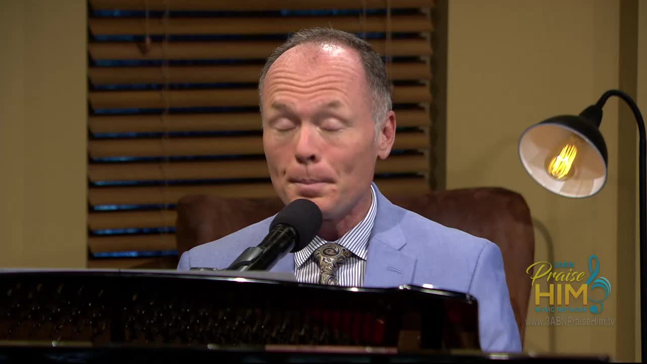 The man, eyes closed, is singing into a microphone. He's wearing a light blue suit jacket, and the "Praise Him" logo is visible in the corner.
