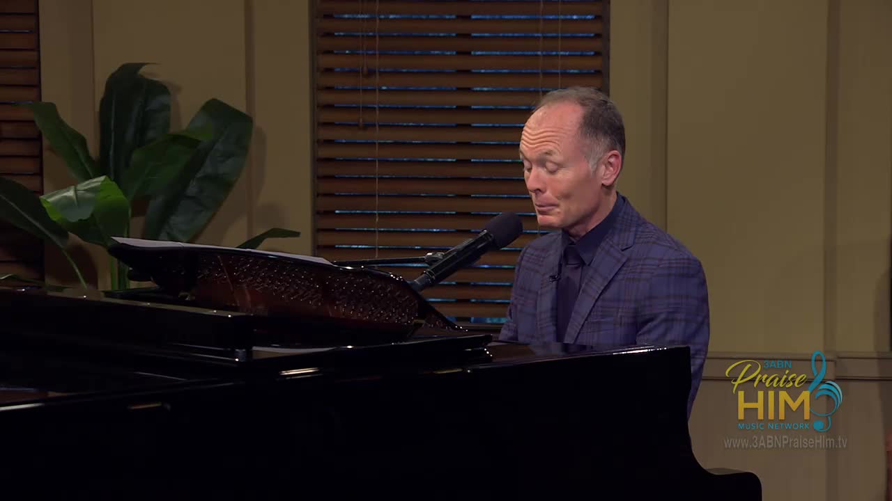 A man in a patterned suit sits at a grand piano, his fingers poised above the keys. He's singing into a microphone, reading music from the stand.
