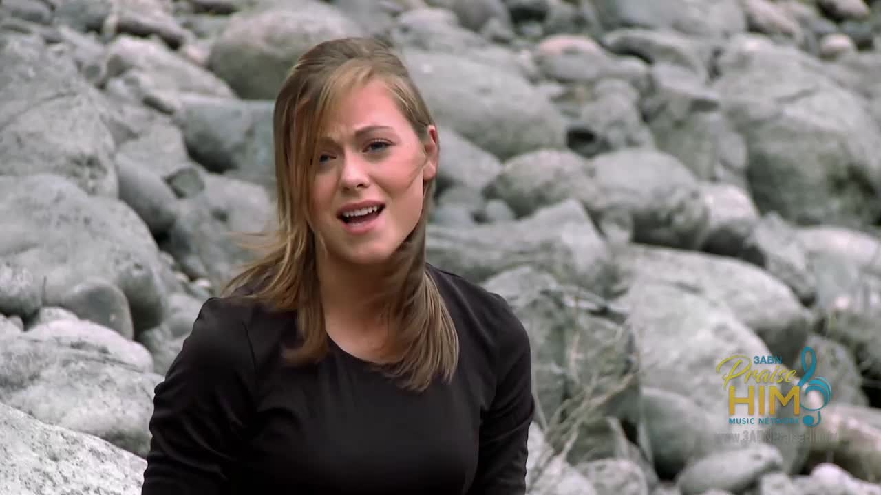 A woman sings, her hair slightly blowing in the breeze. Behind her, a pile of large, gray rocks forms a backdrop.
