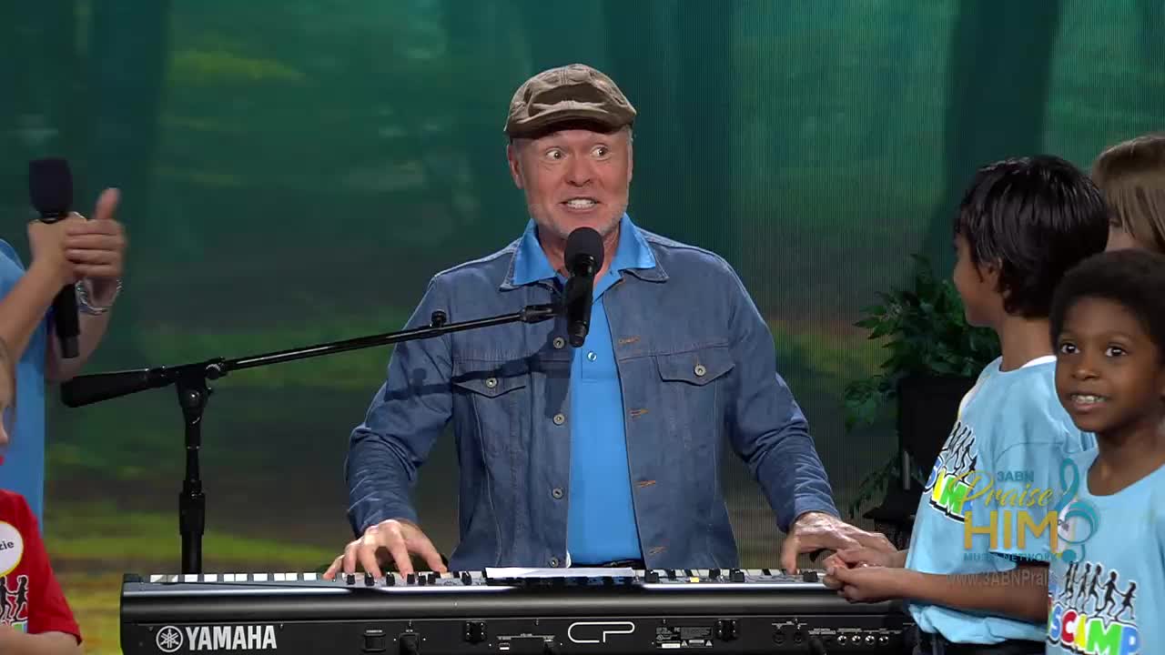 The man in the cap plays the keyboard, his face animated as he sings into the microphone. Children in matching blue shirts stand beside him, some smiling and others looking on with interest.
