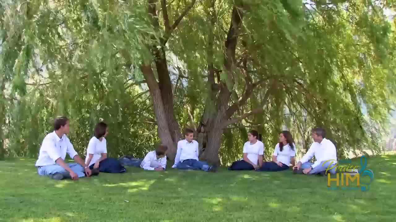 Seven people sit in a circle on the grass beneath a large, weeping willow tree. They are all dressed in white shirts and dark pants, and the "Praise Him" logo is visible in the corner.
