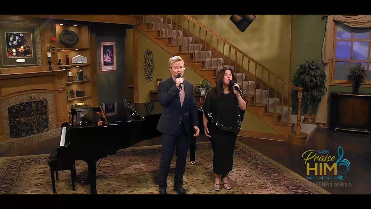 A man and a woman, each holding a microphone, stand on a patterned rug, singing. Behind them, a grand piano sits near a fireplace, and a staircase leads upwards.
