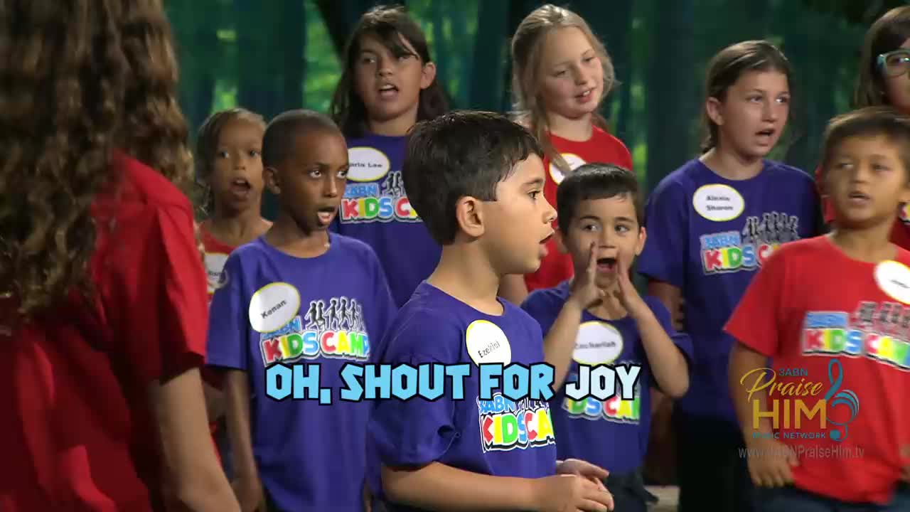 A group of children in matching shirts are singing, their mouths wide open. The words "OH, SHOUT FOR JOY" appear on the screen, likely during a performance on the 3ABN Praise Him Music Network.
