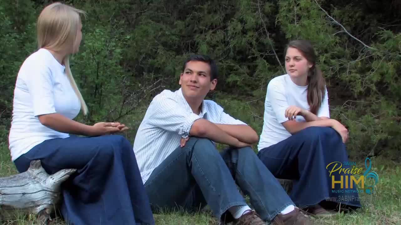 Three people, dressed in white shirts and denim skirts or pants, sit on a log and the ground. One man in a button-down shirt looks up with a slight smile.
