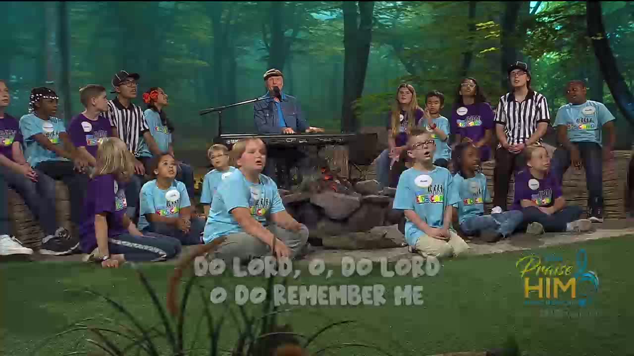 A group of children and an adult are singing, with the words "Do Lord, O, Do Lord, O Do Remember Me" on the screen. The man at the keyboard is wearing a denim jacket.
