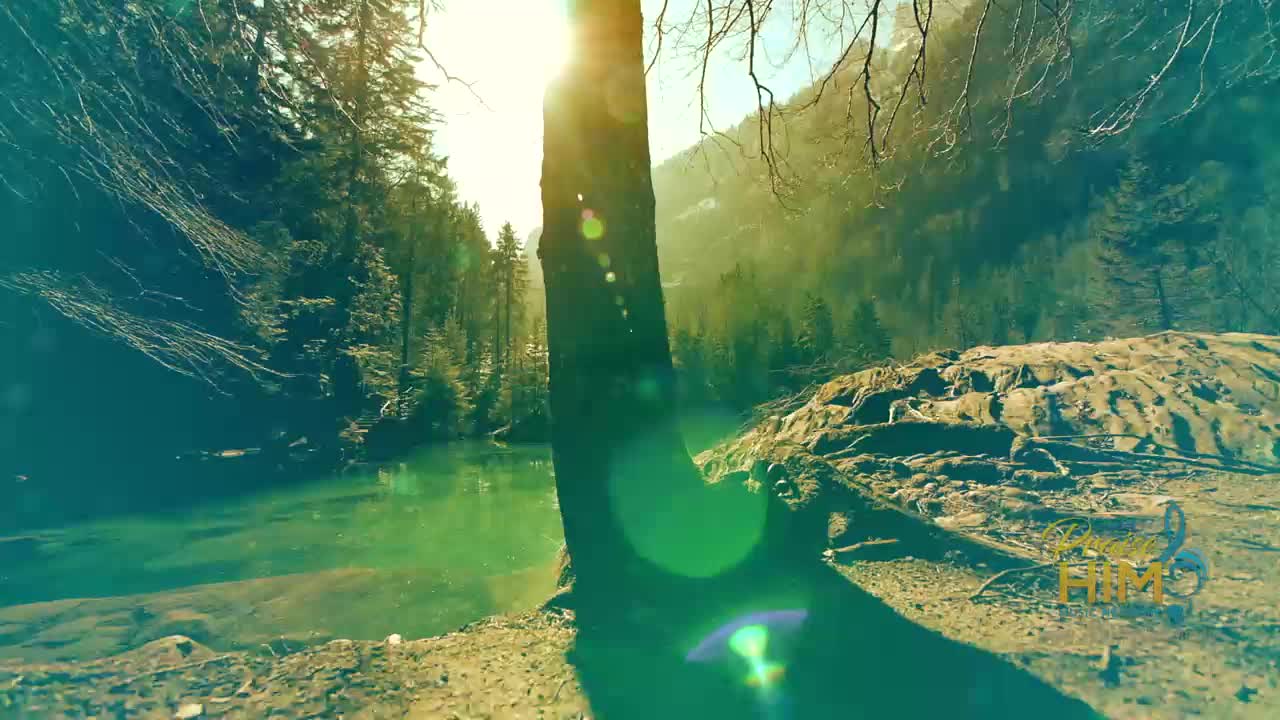 Sunlight streams through the trees, illuminating a still, turquoise lake. A tree trunk casts a dark shadow across the foreground, while the 3ABN Praise Him Music Network logo is visible in the corner.

