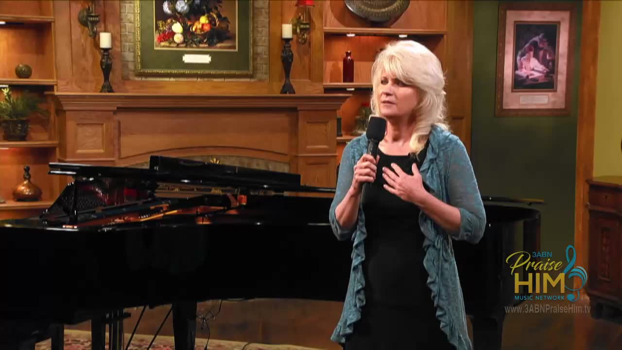 A woman with blonde hair holds a microphone, her hand resting on her chest. Behind her, a grand piano gleams under the lights of a set for 3ABN Praise Him Music Network.

