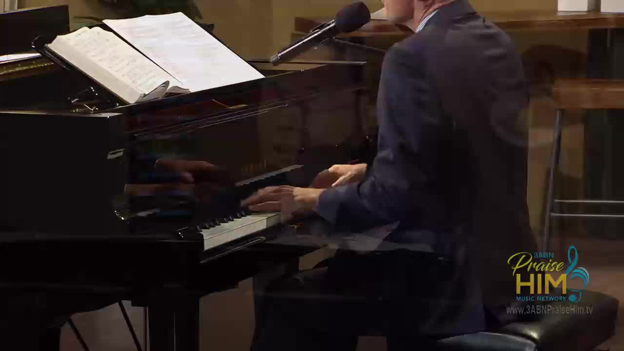 A man in a dark suit plays the piano, his fingers dancing across the keys.  Sheet music sits open on the stand, and a microphone hovers nearby.
