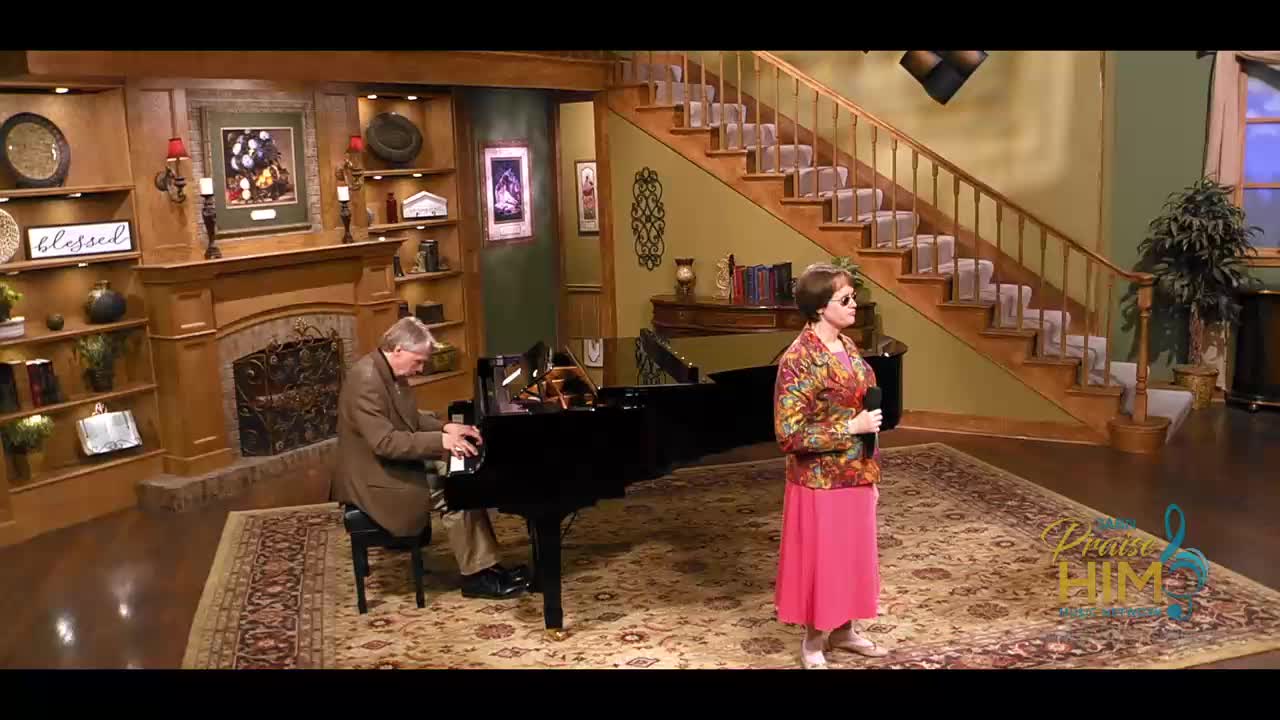 A man in a brown suit plays the piano, his fingers moving across the keys. A woman in a floral top and pink skirt stands beside him, holding a microphone.
