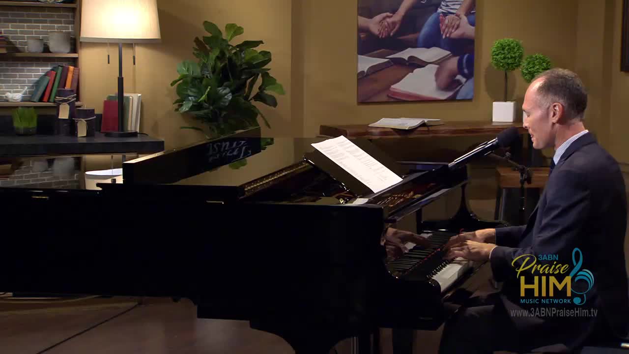 A man in a suit sits at a grand piano, his fingers dancing across the keys. He's singing into a microphone, with sheet music propped up in front of him.
