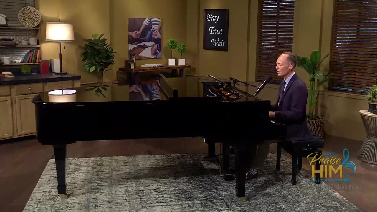 A man in a dark suit sits at a grand piano, playing and singing into a microphone. The scene is set in what looks like a studio for the 3ABN Praise Him Music Network in the United States, with a cozy, homey backdrop.
