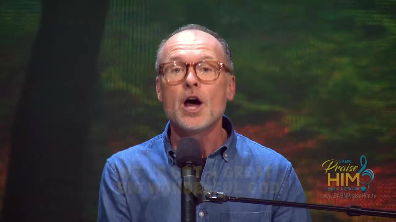 A man with glasses, wearing a denim shirt, speaks into a microphone. Behind him, a backdrop shows a blurred scene, and the 3ABN Praise Him Music Network logo is visible on the right.
