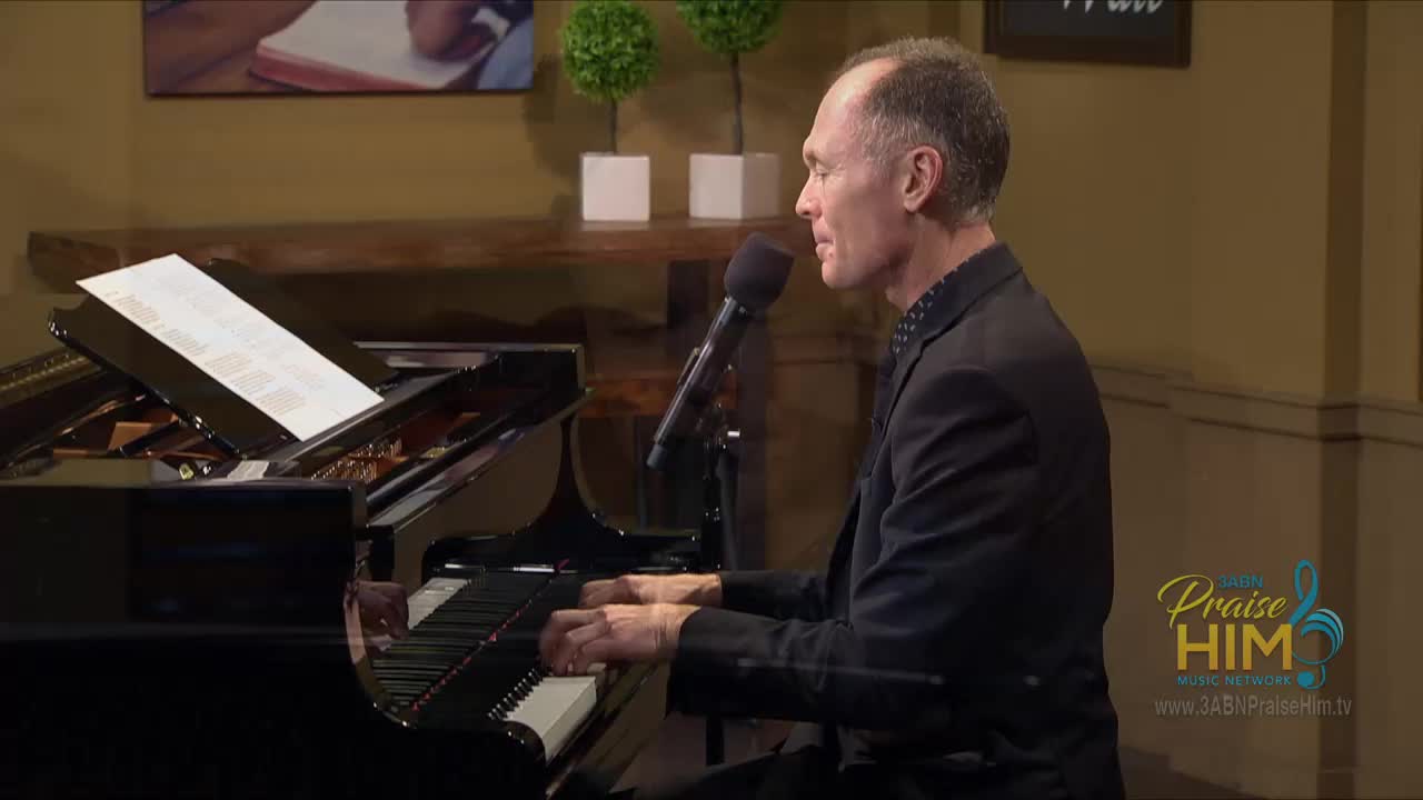 A man in a black suit sits at a grand piano, his fingers moving over the keys. He sings into a microphone, the music sheets reflecting the light from the studio.

