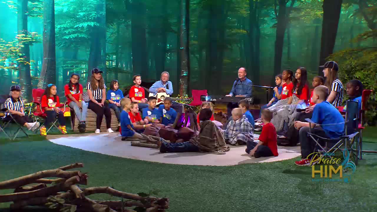 A group of children and adults are gathered around a pretend campfire, with two men playing instruments. The scene is part of a program from the 3ABN Praise Him Music Network, filmed in the United States, with a forest backdrop.
