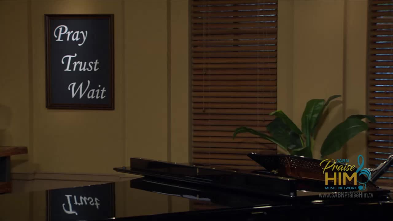 A sign on the wall reads "Pray, Trust, Wait" in elegant script. The glossy black surface of a piano reflects the words, along with the logo for 3ABN Praise Him Music Network.
