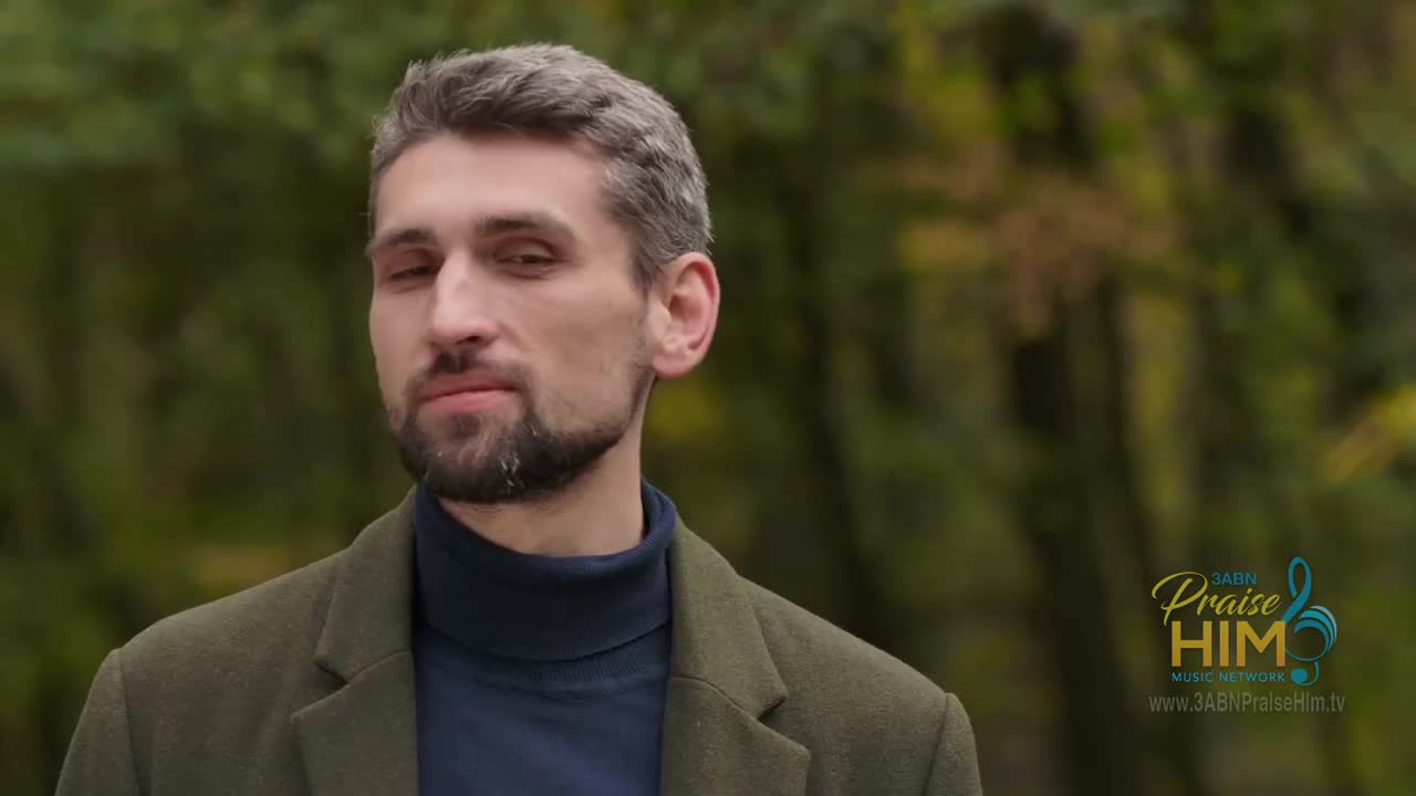 A man with graying hair and a trimmed beard stands in front of a blurred background of trees. He's wearing a dark turtleneck and a green jacket, and the logo for 3ABN Praise Him Music Network is visible in the corner.
