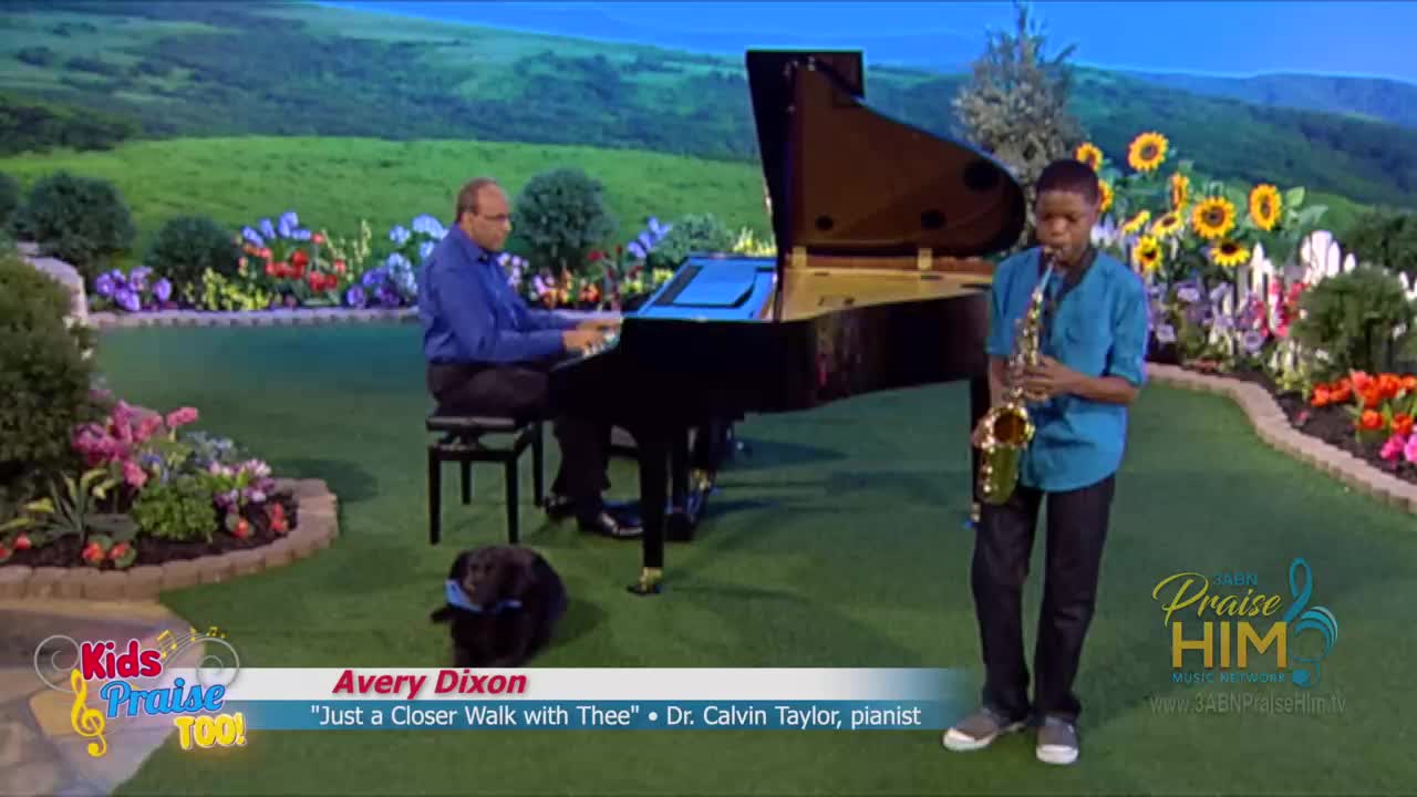 A young man in a blue shirt stands with a saxophone, facing the camera. A man in a blue shirt plays the piano, while a small dog sits nearby, all on a set for 3ABN Praise Him Music Network.
