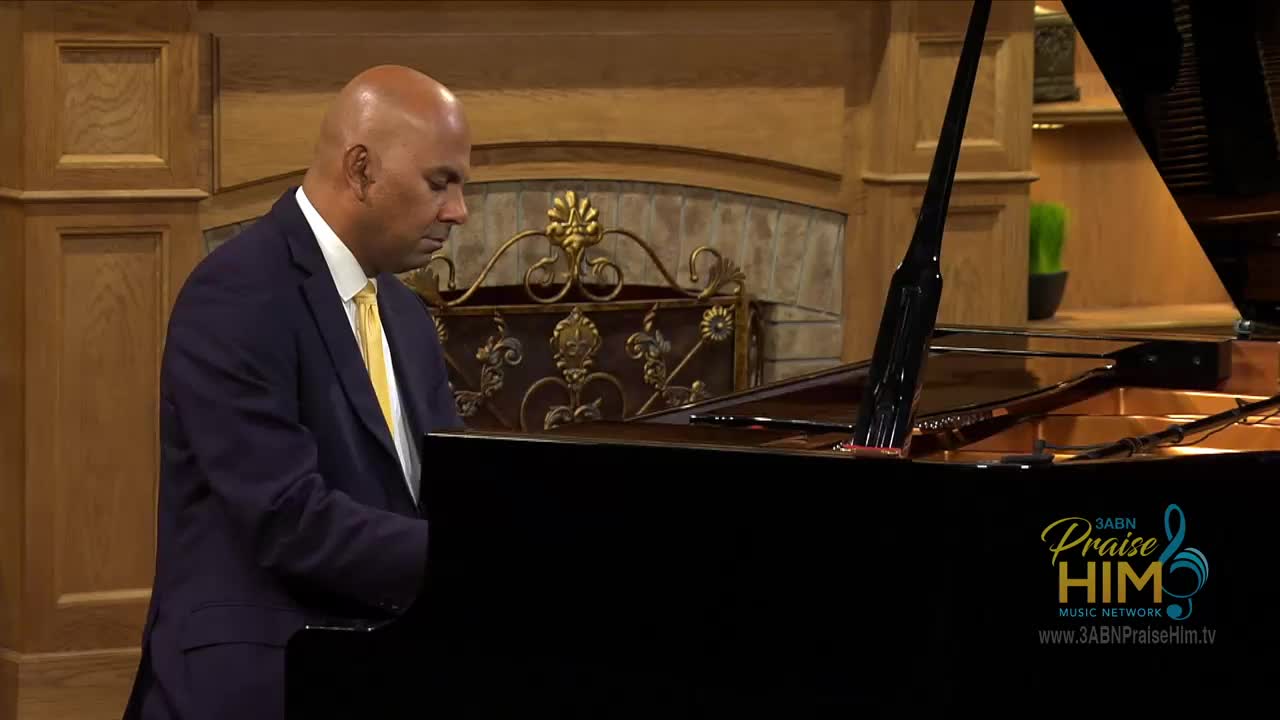 A man in a navy suit and yellow tie plays the piano, his fingers moving across the keys. The 3ABN Praise Him Music Network logo is visible in the corner.
