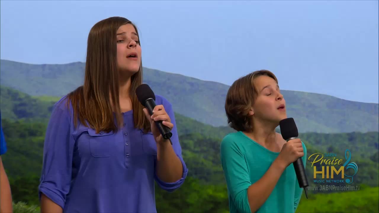 Two young women stand close together, each holding a microphone and singing with their eyes closed. Behind them, a backdrop shows a painted landscape, while the 3ABN Praise Him Music Network logo is visible in the corner.
