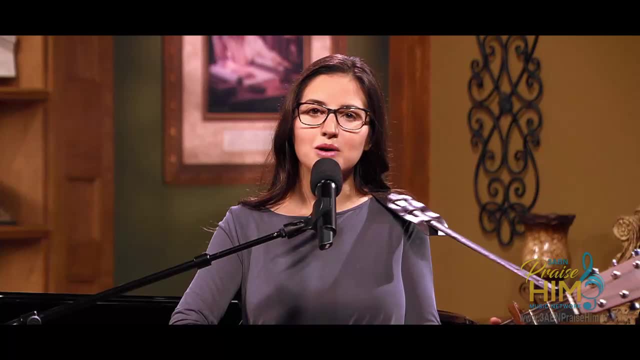 A woman with glasses stands before a microphone, singing, with a guitar resting on her shoulder. The scene is part of a musical performance on the 3ABN Praise Him Music Network, filmed in the United States.
