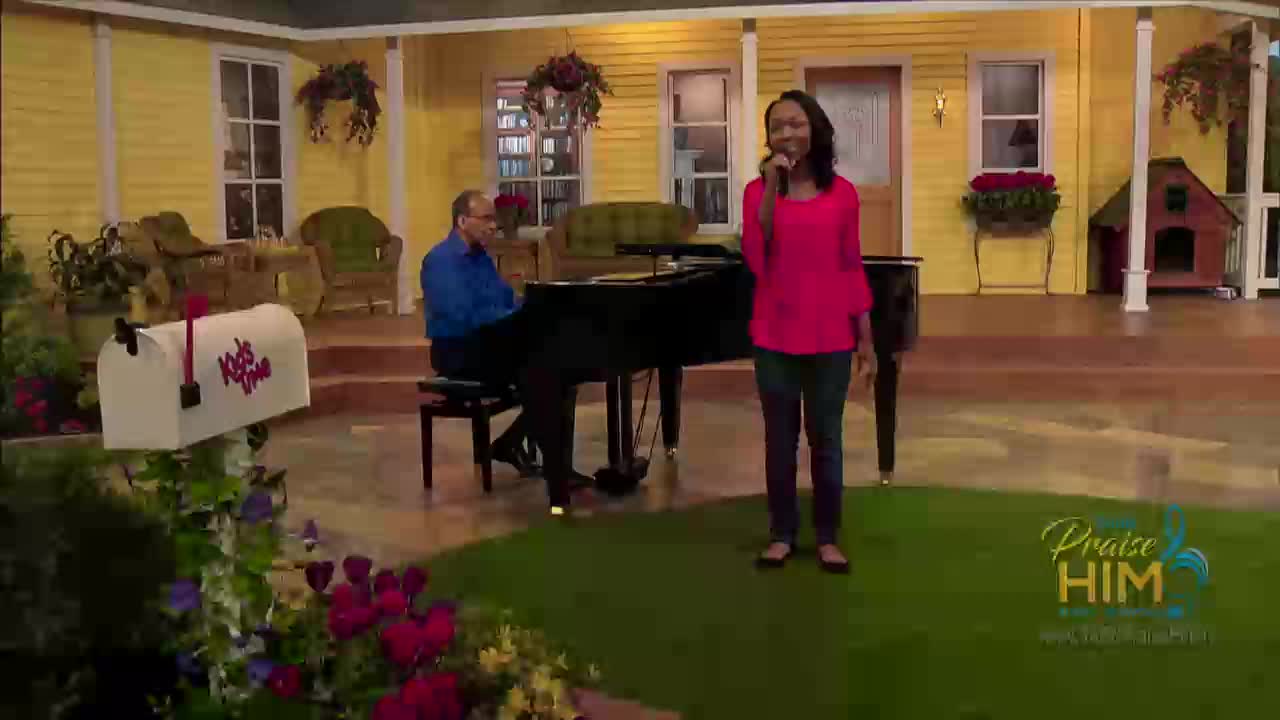 A woman in a bright pink top sings into a microphone on a stage designed to look like a front yard. Behind her, a man in a blue shirt plays a grand piano.

