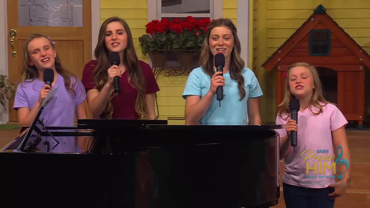 Four young women stand behind a grand piano, each holding a microphone. They are singing, their faces animated as they perform on the 3ABN Praise Him Music Network set.
