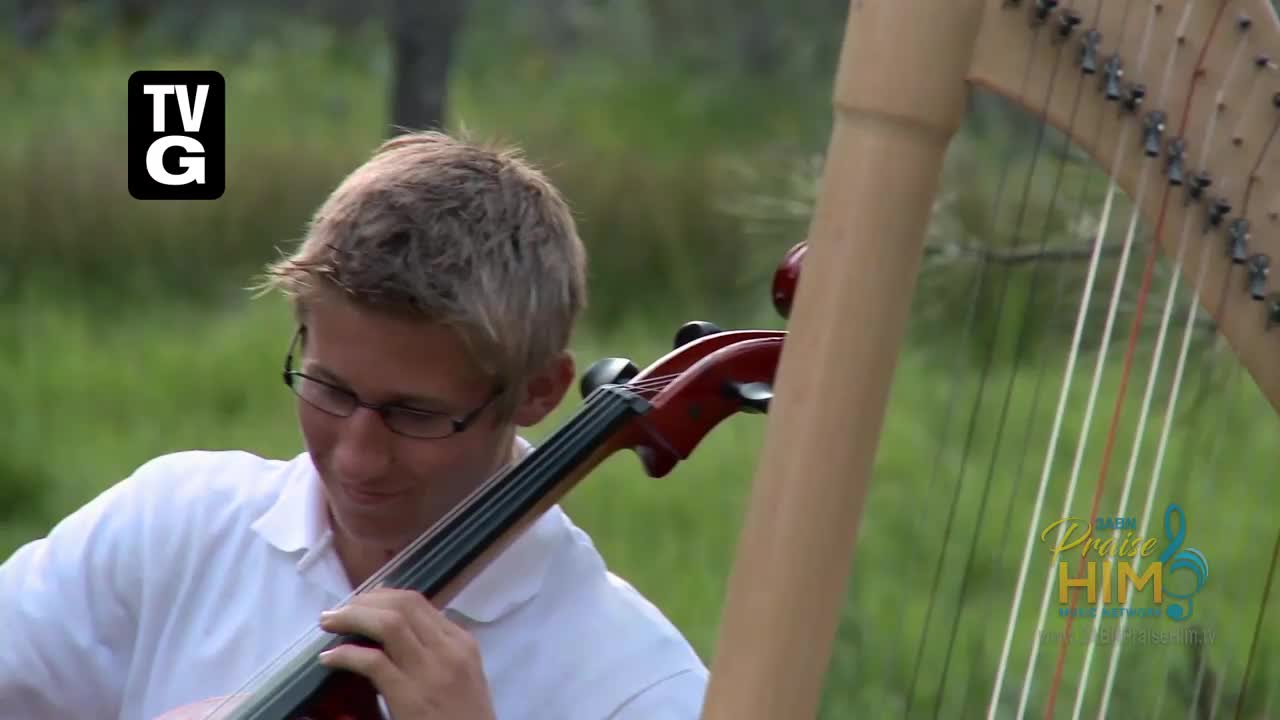 A young man in glasses is playing a cello, his fingers moving on the strings. Beside him, a harp stands tall against a backdrop of green foliage. 
