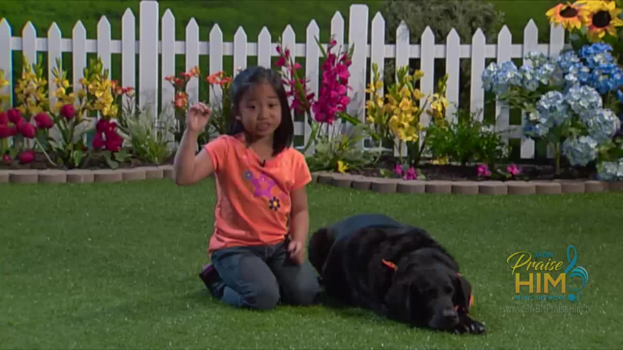 A young girl kneels on the grass, gesturing with her right hand. Beside her, a black dog rests, its head down, in a garden setting with a white picket fence and colorful flowers.
