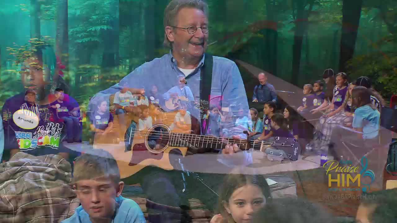 A man with glasses strums an acoustic guitar, smiling at the camera. Children in matching shirts sit nearby, and the "Praise Him" logo is visible on the lower right.
