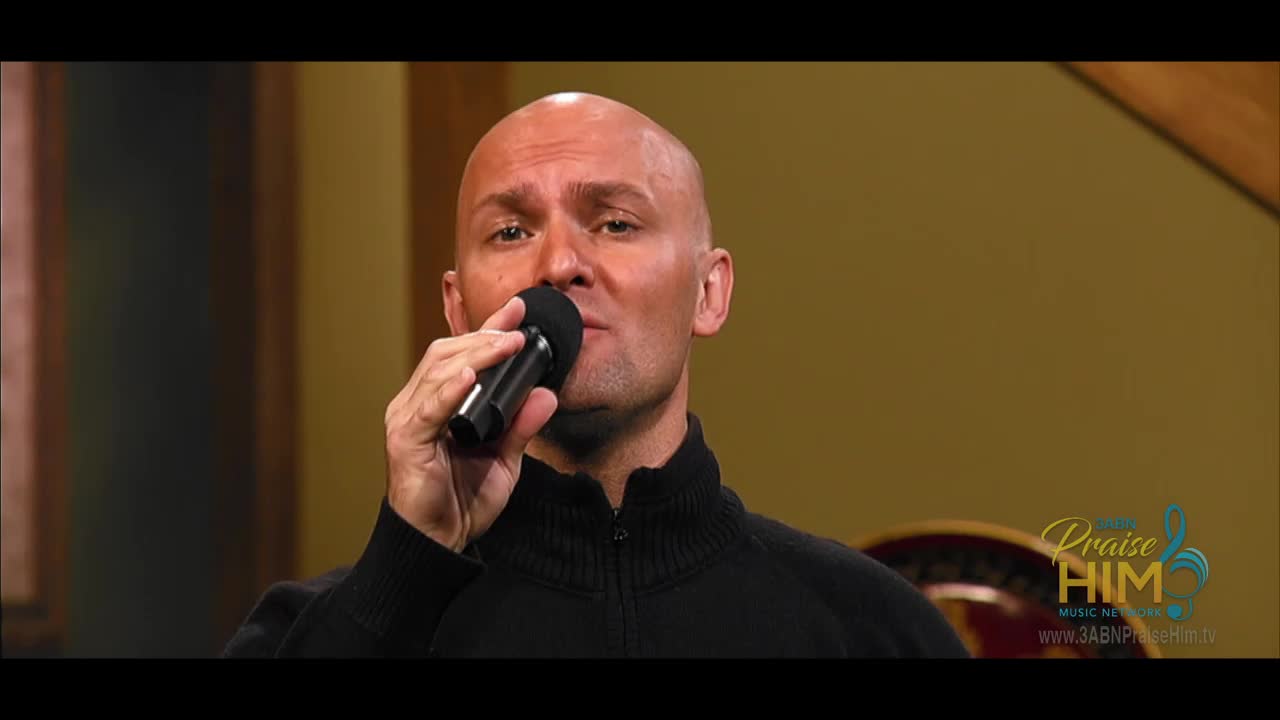 A man holds a microphone up to his mouth, singing on the 3ABN Praise Him Music Network. He's wearing a black sweater in a studio setting.

