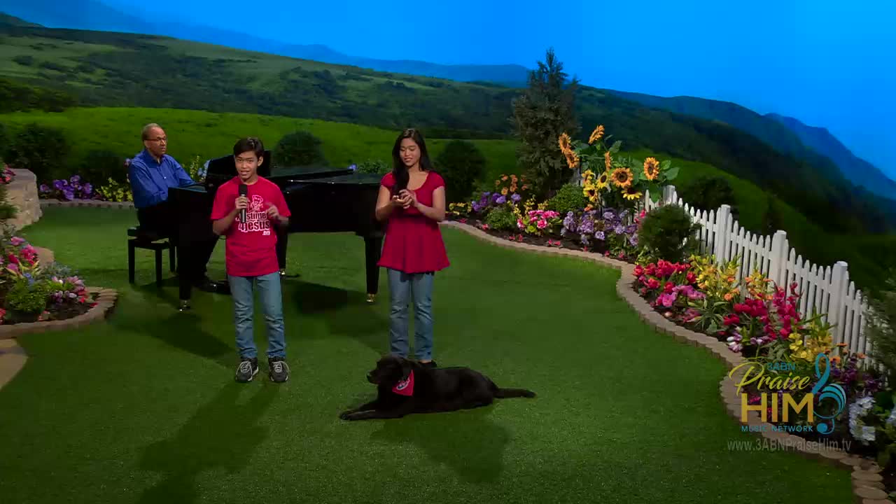 A young boy sings into a microphone while a woman stands beside him, holding a small device. Behind them, a man plays the piano, and a black dog rests on the artificial grass.
