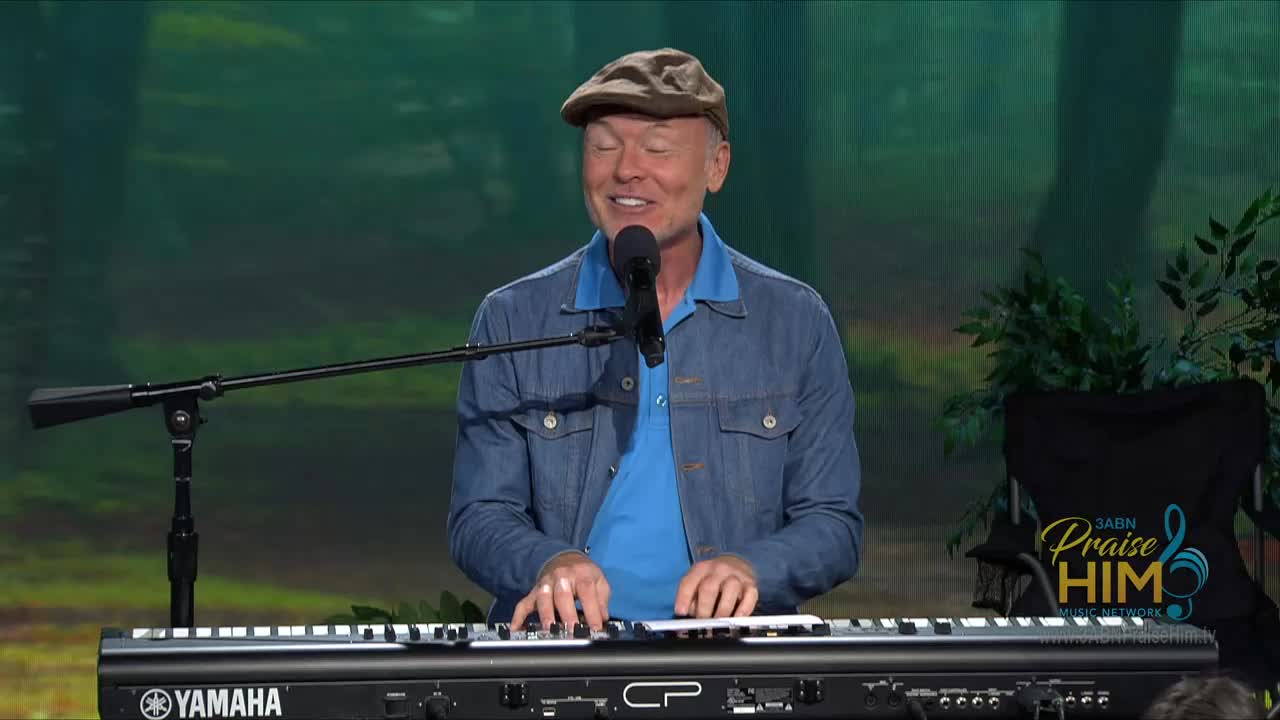 The man in the denim jacket and cap is smiling as he plays the keyboard. He's on the 3ABN Praise Him Music Network stage in the United States, with a backdrop that looks like a forest.
