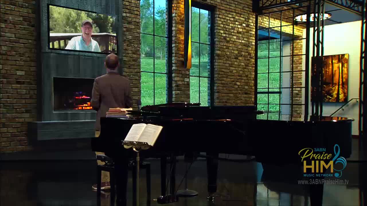 A man in a suit stands facing a grand piano, with sheet music open on the stand. On the screen above the fireplace, a man is speaking, likely for the 3ABN Praise Him Music Network.
