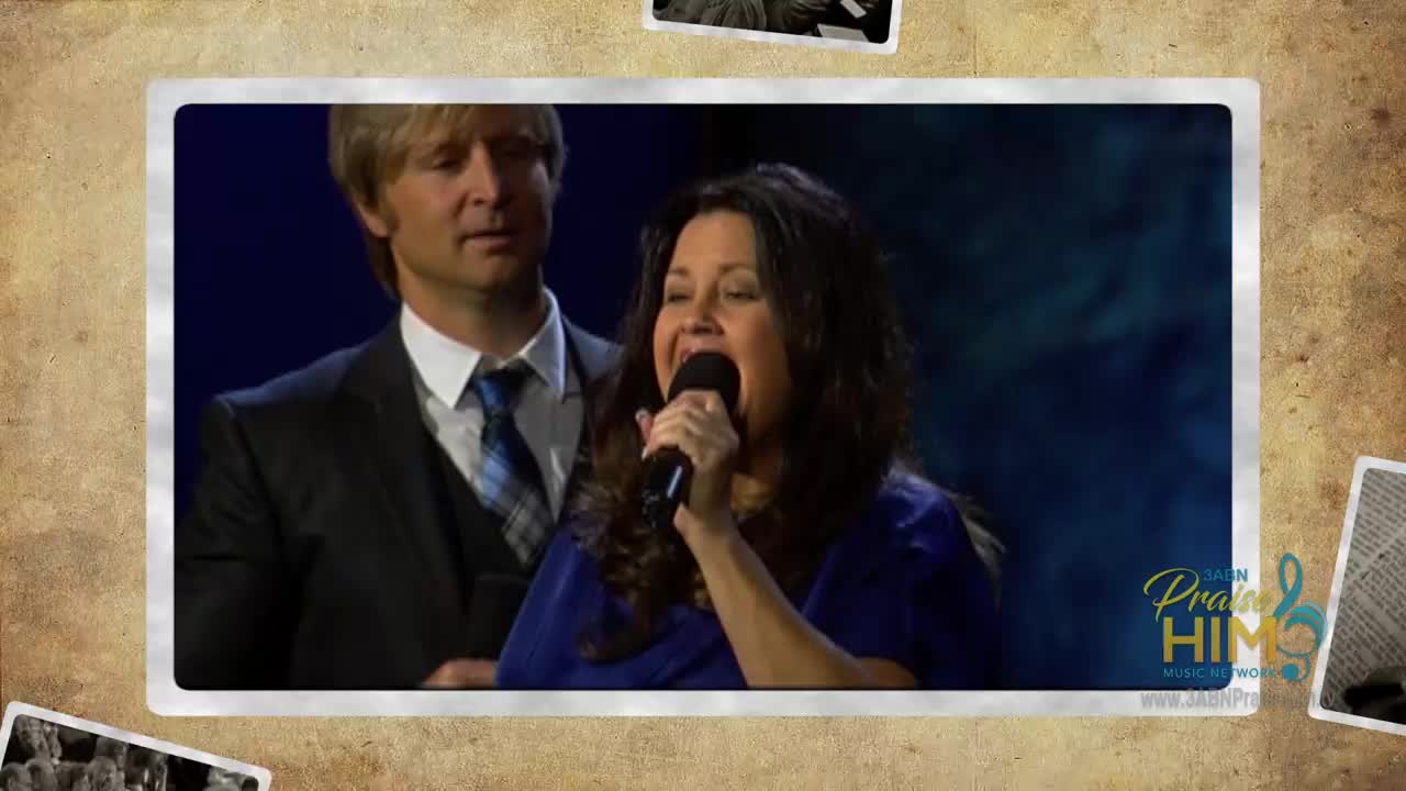 A woman in a blue dress belts into a microphone, her face lit up. Behind her, a man in a suit looks on, part of a broadcast for 3ABN Praise Him Music Network.
