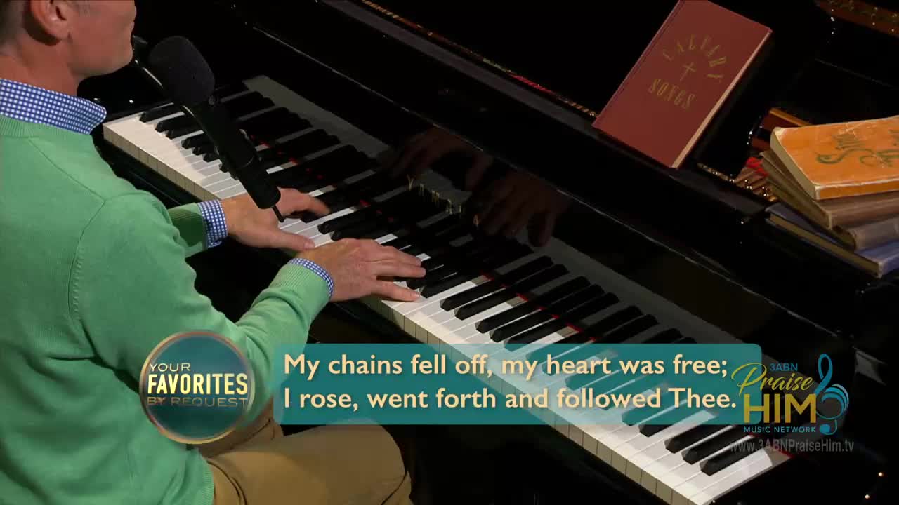 A man in a green sweater plays the piano, his hands moving over the keys. Beside him, a microphone stands ready, and a hymn book rests open on the piano's surface.

