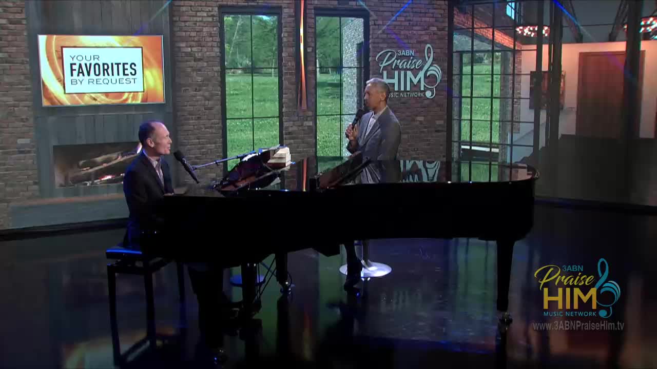 A man in a suit sits at a grand piano, facing a microphone. Across from him, another man holds a microphone and speaks on the set of 3ABN Praise Him Music Network.
