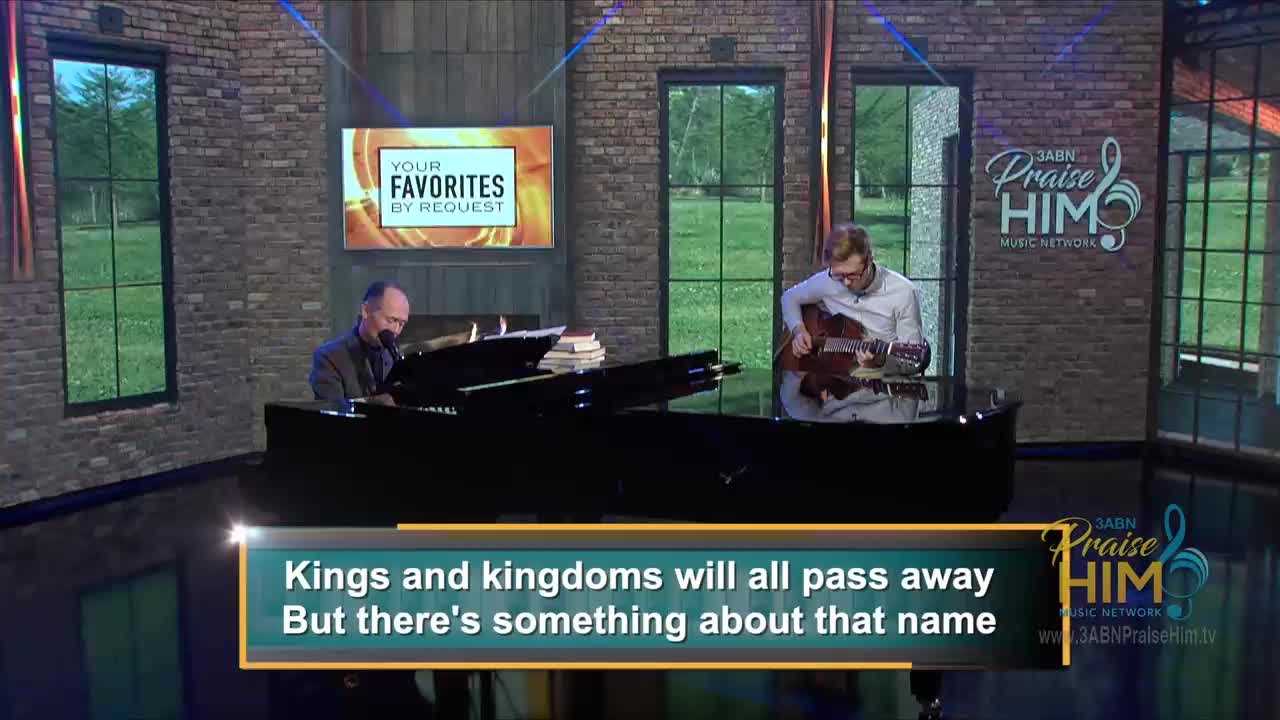 A man in a suit plays the piano, his fingers dancing across the keys. Beside him, another man strums a guitar, both musicians illuminated on a set for 3ABN Praise Him Music Network.
