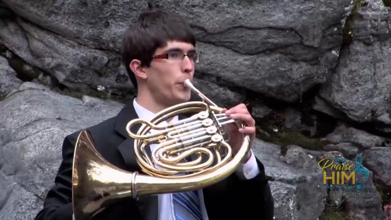 A young man in a suit is playing a French horn against a backdrop of gray rocks. He's performing for the 3ABN Praise Him Music Network, a broadcast from the United States.
