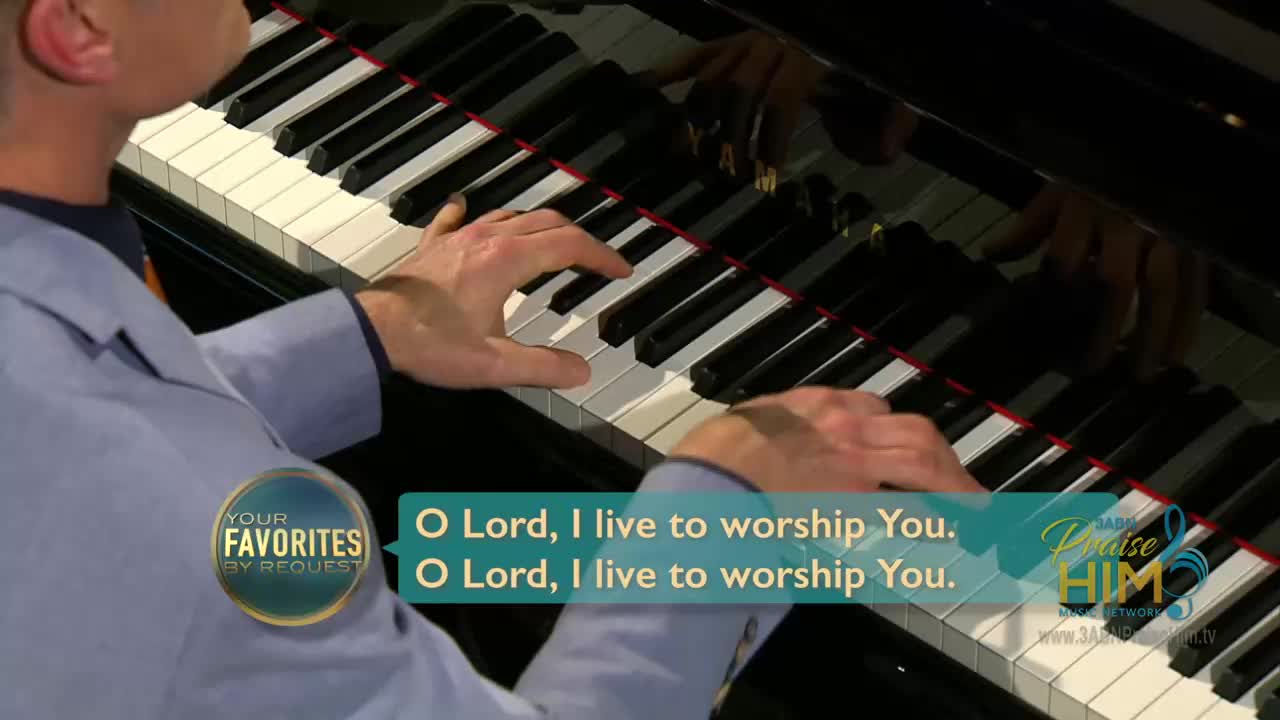 A man in a light blue jacket plays the piano, his fingers dancing over the keys. The words "O Lord, I live to worship You" are displayed on the screen, a segment from 3ABN Praise Him Music Network.
