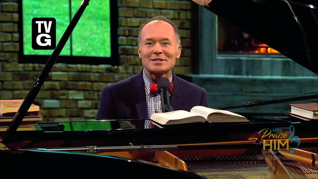 A man in a suit and bow tie sits at a grand piano, smiling at the camera. He's on the set of 3ABN Praise Him Music Network, with a book open in front of him.
