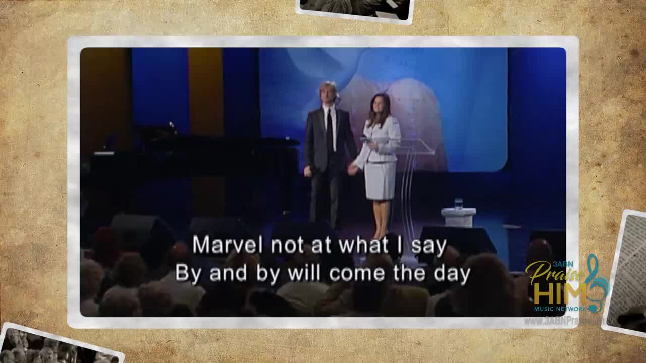 A man in a dark suit stands next to a woman in a white suit, both facing the audience. The woman is at a podium, and text about the day appears below them.
