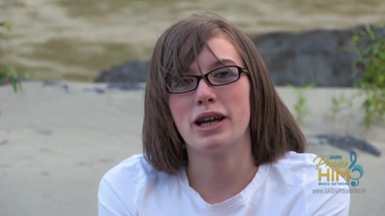 A young woman with glasses speaks directly to the camera, likely for the 3ABN Praise Him Music Network. The background shows a sandy area with some greenery.
