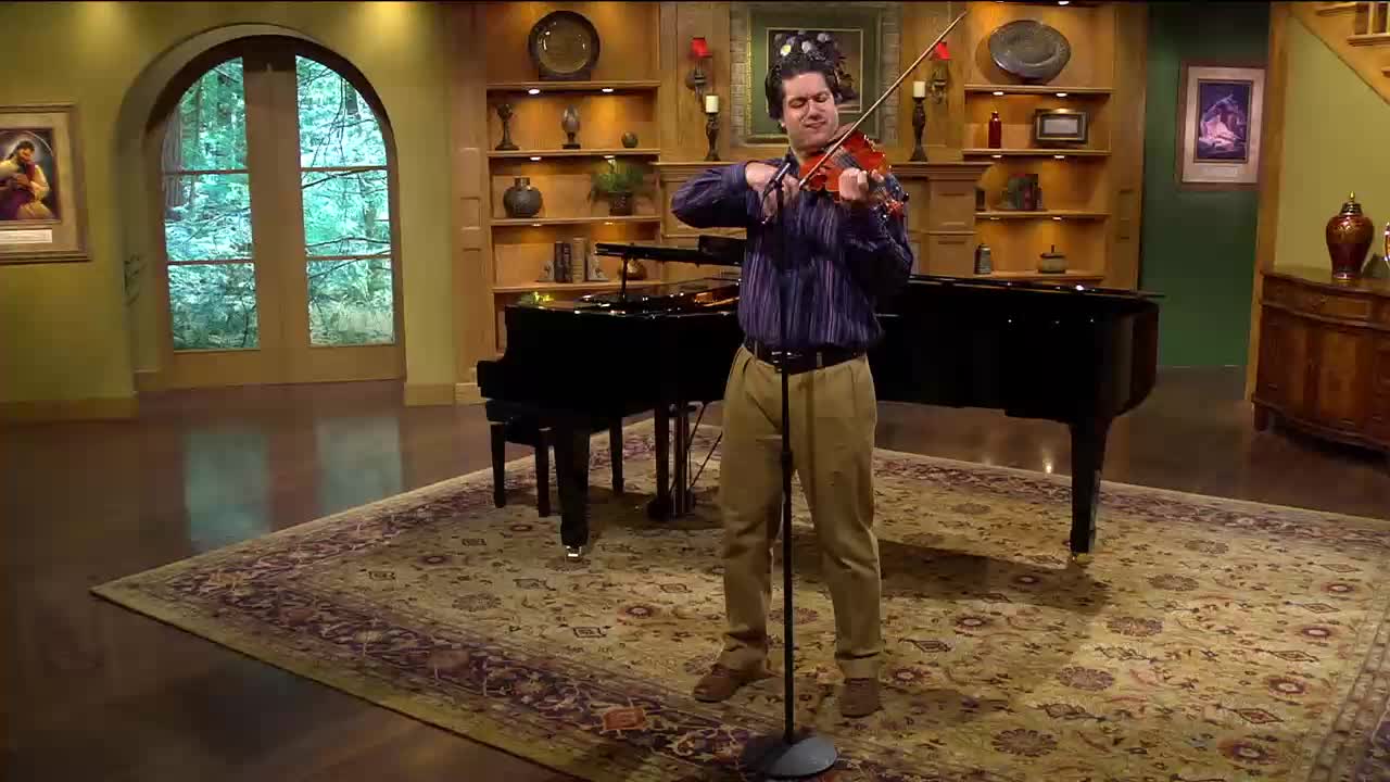 A man stands playing a violin in front of a grand piano, his bow moving across the strings. He's in a room with warm lighting and decorative shelves.