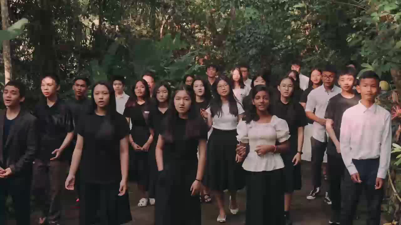 A group of young people in dark clothing stands in a lush, green outdoor setting, their faces turned forward as if singing. They are part of a choir, likely preparing for a performance or recording for a program like 3ABN Praise Him Music Network.