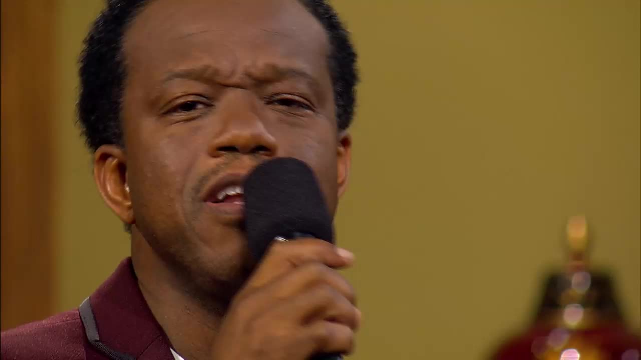 A man sings passionately into a microphone, his brow furrowed in concentration. He's wearing a deep red jacket, and behind him, a golden wall is visible with a blurred ornament.