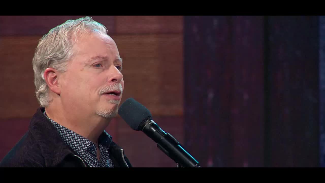 A man with graying hair and a mustache sings into a microphone. He wears a dark jacket over a patterned shirt.