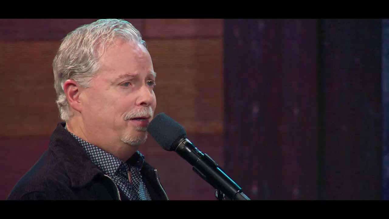 A man with graying hair and a mustache sings into a microphone. He wears a dark jacket over a patterned shirt, likely on a program for the 3ABN Praise Him Music Network.