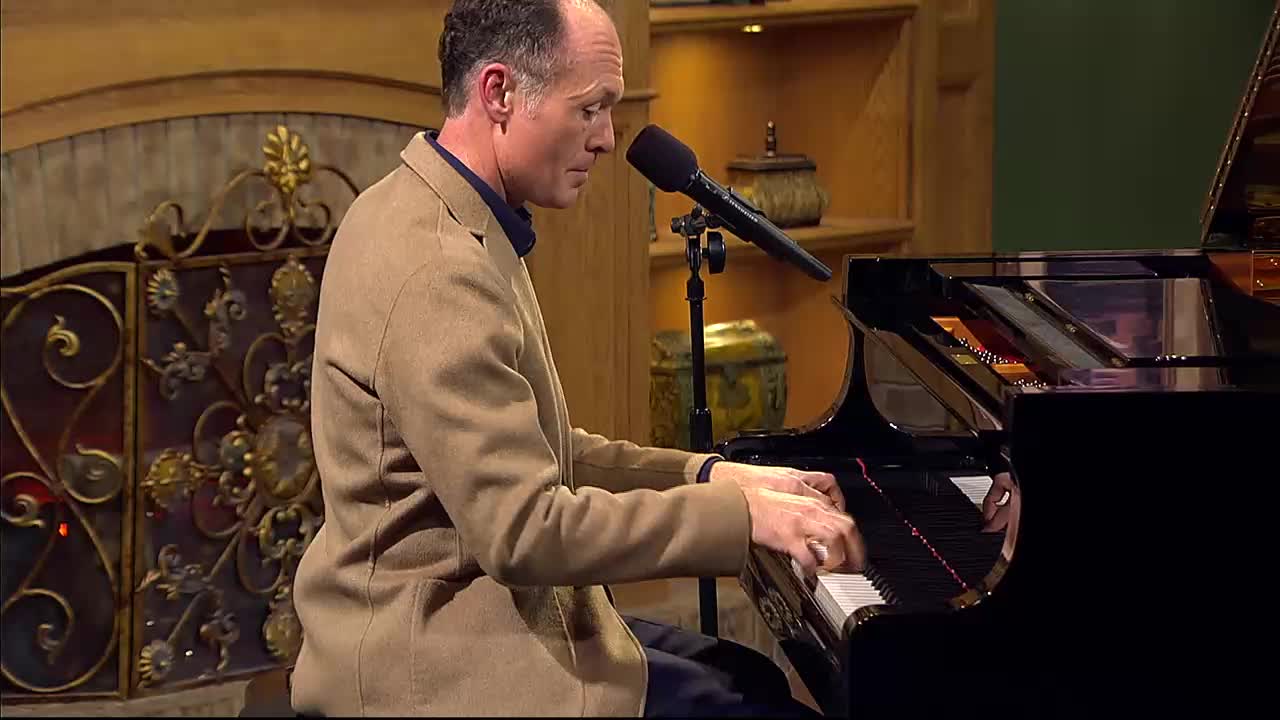 A man in a tan jacket plays a grand piano, his fingers dancing across the keys. He's performing in a studio setting, likely for a broadcast on the 3ABN Praise Him Music Network.